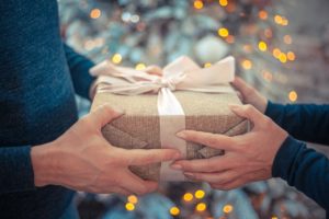 Two people exchange a gift wrapped in brown paper with a light pink ribbon, with a blurred, festive background featuring warm, glowing lights.