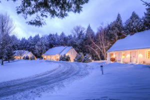 Several cozy white houses with warm glowing windows are nestled among snow-covered trees and a winding snowy road at dusk, evoking the peaceful charm of Vermont luxury hotel accommodations in a tranquil winter scene.