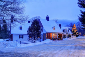 Snow-covered houses with warm glowing windows evoke the charm of Vermont luxury hotel accommodations, as a lit Christmas tree stands outside in a peaceful winter landscape at dusk, blue sky fading above snowy rooftops and ground.