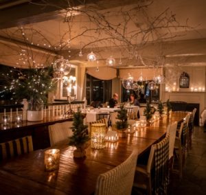 A cozy, warmly lit restaurant in Stowe, Vermont with a long wooden table decorated with candles, small pine trees, and hanging glass ornaments sets the scene for an intimate Thanksgiving dinner. Two people enjoy a quiet meal in the background.
