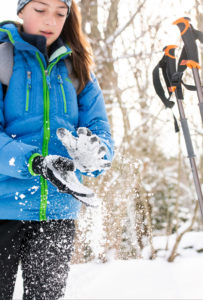 A person in a blue winter jacket and gloves is clapping their hands, causing snow to scatter. They stand outdoors with ski poles in the snow, ready for cross country skiing among trees on a sunny winter day.