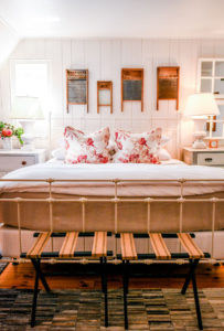 A cozy bedroom with a white metal bed, floral pillows, two bedside tables with lamps, vintage washboards on the wall, and a wooden bench at the foot of the bed. Many decor pieces were acquired through donation, adding to its bright and inviting charm.