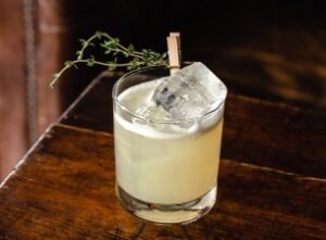 A cocktail in a clear glass, filled with a pale yellow liquid and ice cubes, garnished with a sprig of herbs held by a small clothespin, sits on a wooden surface—perfect for savoring the fine dining experience in Stowe, VT.