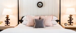 A neatly made bed with white linens, blush pink pillows, and a smaller gray pillow creates a cozy retreat. Modern lamps on wooden nightstands and a round mirror above the bed evoke the inviting charm of unique hotels in Vermont.