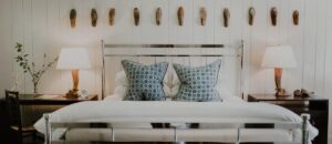 A cozy Edson Hill Guest Rooms bedroom with a metal-framed bed, blue patterned pillows, white bedding, two wooden bedside tables with lamps, and wood shoe molds mounted on a white paneled wall above the bed.
