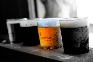 Four plastic cups filled with different types of beer, ranging in color from light amber to dark brown, are lined up on a wooden surface with chalk markings.