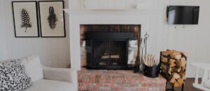 A cozy living room, inspired by Edson Hill Guest Rooms, features a brick fireplace, stacked firewood, fireplace tools, a mounted TV, and framed feather artwork. A white couch with patterned pillows sits invitingly on the left.