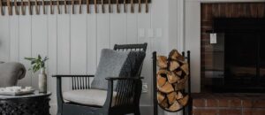 A cozy living room, inspired by Edson Hill Guest Rooms, features a black armchair with gray cushions, a round table with a vase of greenery, stacked firewood, and a brick fireplace against a white-paneled wall with wooden accents.
