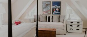A cozy attic bedroom inspired by Edson Hill Guest Rooms features a white bed, a couch with gray pillows, a white nightstand, and two framed ski-themed posters. Light streams in from a triangular window.