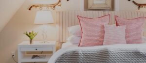 Cozy Edson Hill Guest Room with a bed featuring red and white patterned pillows, a striped headboard, a white nightstand with a lamp, and a small vase of flowers. Walls adorned with rustic wooden accents create an inviting atmosphere.