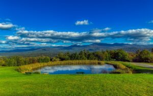 A small pond surrounded by green grass and trees, with benches nearby. Rolling hills and distant mountains are visible under a blue sky with scattered clouds.