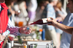 A person pours red wine from a bottle into a glass held by another person at an outdoor event, with others and refreshments visible in the blurred background.