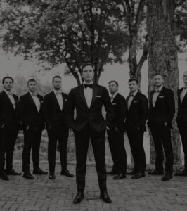 A group of eight men in tuxedos stand outdoors on a stone path, with one man in the center slightly ahead of the others, all posing confidently among trees.