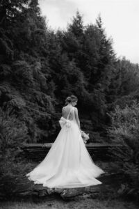 A bride in a flowing white gown stands with her back to the camera, holding a bouquet and facing a dense forest of tall trees—an elegant black and white moment from the Gretchen & Chris Edson Hill Wedding.