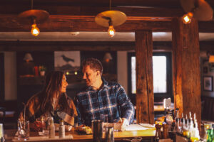 A man and woman sit together at a cozy bar, warmly lit by hanging bulbs. They smile over drinks and chat about weekend packages, surrounded by rustic wooden beams and shelves in the background.