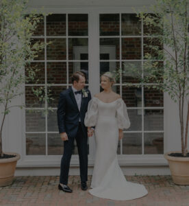 A bride in an off-the-shoulder white gown and a groom in a black tuxedo hold hands and smile at each other, standing on a brick patio in front of large glass windows framed by potted plants.