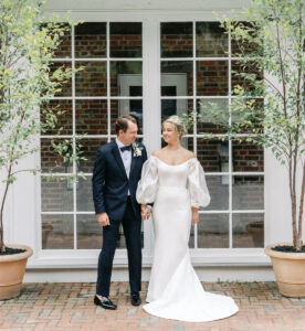 A bride in an off-the-shoulder white gown with puffed sleeves stands holding hands with a groom in a black tuxedo, both smiling in front of large windows and two potted trees.