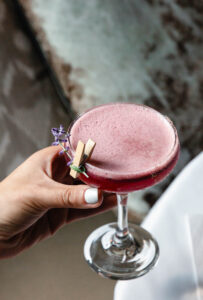 A hand with white nail polish holds a coupe glass filled with a frothy pink cocktail from the menu, garnished with a small sprig of purple flowers and a tiny clothespin.