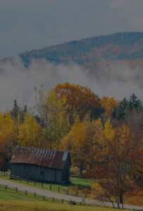 A rustic wooden barn sits among colorful autumn trees at Edson Hill - Stowe Vermont Inn, with rolling hills and mist rising in the background.