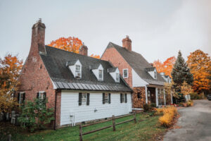 A large, two-story manor house with brick and white siding, steep rooflines, and multiple chimneys evokes the charm of Vermont luxury hotel accommodations, surrounded by trees with vibrant autumn foliage under a cloudy sky.