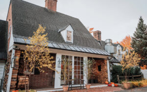 A cozy brick house with a steep roof, white trim, and large windows, reminiscent of Edson Hill - Stowe Vermont Inn, decorated with autumn trees, pumpkins, string lights, and surrounded by colorful fall foliage.