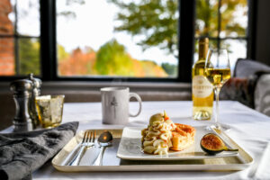 A table set for one in a casual fine dining style, with fork, spoon, and knife, features a dessert topped with cream and fig. Nearby, a mug, wine bottle, and glass complement the scenic window view of trees in the background.