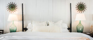 A neatly made bed with white bedding and pillows sits against a white paneled wall in the inviting style of Edson Hill Rooms, flanked by green glass lamps with white shades and decorative sunburst wall hangings above each lamp.