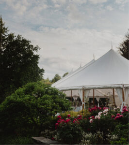 A large white event tent stands in a lush garden with blooming pink and white flowers in the foreground and green trees under a cloudy sky.