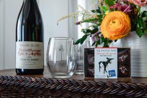 A bottle of red wine, two empty glasses, a box of farmstead milk caramels, and a colorful floral arrangement are displayed on a woven surface—perfect for celebrating Reservation Enhancements in style.