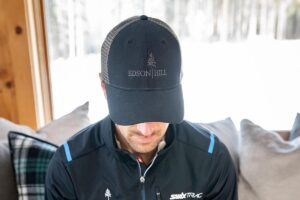 A man wearing a dark baseball cap with EDSON HILL and a tree logo, sitting indoors by a window on a couch with plaid pillows, looks downward while considering recent Reservation Enhancements.