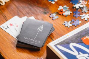 A deck of Edson Hill playing cards and scattered puzzle pieces rest on a wooden table beside a partially visible book, evoking the charm of classic pastimes—perfect for unwinding after experiencing our latest Reservation Enhancements.