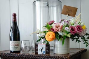 A bottle of red wine, two empty glasses, a box of chocolates, and a colorful bouquet in a white vase are arranged on a woven table. A card peeks from the bouquet—perfect for celebrating Reservation Enhancements in style.
