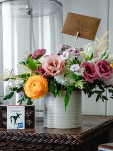 A colorful bouquet of flowers in a white tin can sits on a table next to a box of chocolates, adding charm like thoughtful reservation enhancements. A brown card with a bee design nestles in the bouquet, with a large clear glass jar in the background.