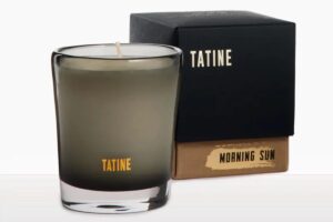 A frosted glass candle labeled TATINE sits next to a black and brown box with TATINE and MORNING SUN written on it in gold lettering, creating an elegant scene reminiscent of thoughtful reservation enhancements.
