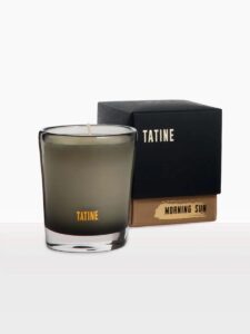 A glass candle with TATINE written on the front sits beside its black and brown box, labeled MORNING SUN. The candle features light-colored wax, a single wick, and evokes a sense of calm—perfect for reflecting on reservation enhancements.