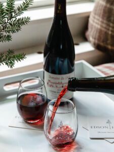 A bottle of red wine sits on a tray beside two glasses, one being filled. The tray holds coasters with a tree design, a small pine branch, and hints at reservation enhancements for an elevated experience.