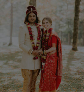 A couple in traditional Indian wedding attire stands outdoors, holding hands and smiling. The groom wears a cream sherwani with a red turban, while the bride wears a red sari with gold details and floral garlands.