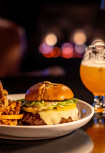 A cheeseburger with lettuce and melted cheese on a white plate next to crispy fries, with a glass of beer in the background and warm, blurred lights—perfect for cozy Tavern Nights at Edson Hill.