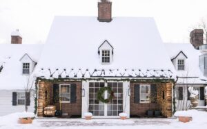 A brick manor with a steep snow-covered roof at Edson Hill - Stowe Vermont Inn, featuring wreath-adorned glass doors, potted evergreens, festive string lights, and deep snow blanketing the grounds and rooftop.