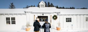 Two people in winter clothing stand with ski poles outside a white building at Edson Hill - Stowe Vermont Inn, adorned with a holiday wreath and potted small trees, surrounded by snow under a clear blue sky.