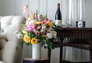 A white vase with a colorful bouquet sits on a small table next to a wicker bar with wine and glasses—perfect for a romantic getaway in Stowe, VT. An upholstered chair rests against the white paneled wall on the left.