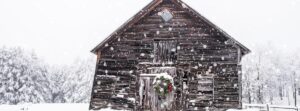 A rustic wooden barn stands in a snowy landscape at Edson Hill - Stowe Vermont Inn, dusted with fresh snow. A festive wreath with red accents hangs on the barn door as large snowflakes fall and snow-covered trees frame the serene scene.