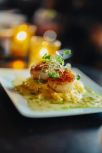 Seared scallops served on a bed of shredded vegetables with a light green sauce, garnished with fresh microgreens, elegantly plated at Edson Hill - Stowe Vermont Inn, with blurred candlelight in the background.