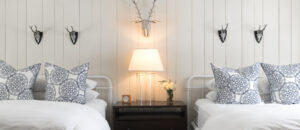 A cozy Edson Hill Guest Rooms bedroom with two white metal-frame beds, each with patterned blue and white pillows. A nightstand with a lamp, clock, and small vase of flowers sits between the beds beneath three deer head wall decorations.