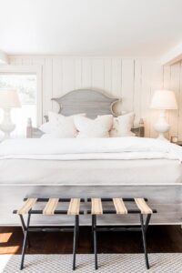 A bright, airy bedroom at Edson Hill - Stowe Vermont Inn features a large bed with white bedding and neutral pillows, flanked by matching bedside tables and lamps. Two wooden stools with woven tops sit at the foot of the bed.