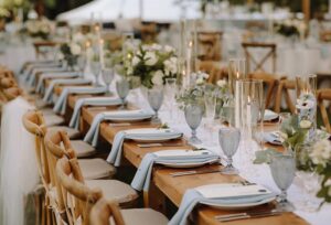 A long wooden table set for an outdoor event at Edson Hill - Stowe Vermont Inn, decorated with white flowers, blue napkins, glass goblets, candles in tall holders, and wooden chairs, creating an elegant and inviting atmosphere.