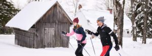 Two people cross-country skiing in the snow past a rustic wooden shed near Edson Hill - Stowe Vermont Inn, both wearing winter sportswear, hats, and sunglasses, with snow covering the ground and rooftops in the background.
