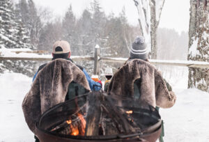 Two people wearing winter hats relax in fur-lined chairs, savoring drinks by a fire pit. Surrounded by snow-covered trees and rustic wooden fencing, they enjoy a cozy retreat—perfect for couples getaway packages.