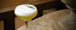 A cocktail from the menu in a coupe glass with a frothy white top, garnished with fresh herbs, sits on a rustic stone surface beside a decorative object.