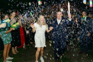A joyful couple, one in a white dress and one in a dark suit, walk hand in hand through a crowd blowing bubbles and waving glow sticks at night, celebrating their elegant wedding at Edson Hill outdoors.